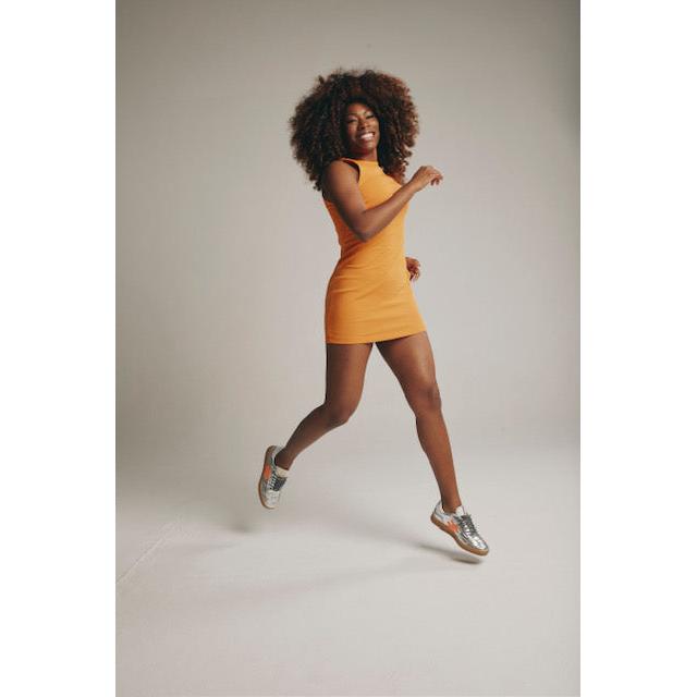Woman wearing metallic silver leather lace-up trainers, orange dress, and playful pose against plain background.