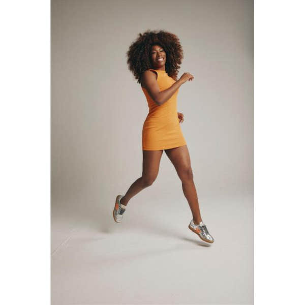 Woman wearing metallic silver leather lace-up trainers, orange dress, and playful pose against plain background.