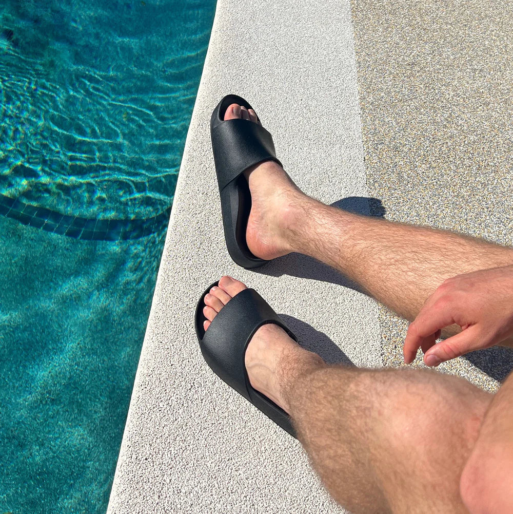 Unisex black waterproof Archies Arch Support Slides on a person's feet by a poolside. Classic slide sandals with arch support.