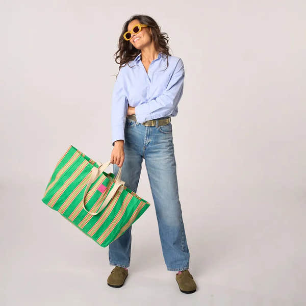 Woman holding Billy Belts 100% organic cotton shopping bag with green stripes, casual outfit, gray background.