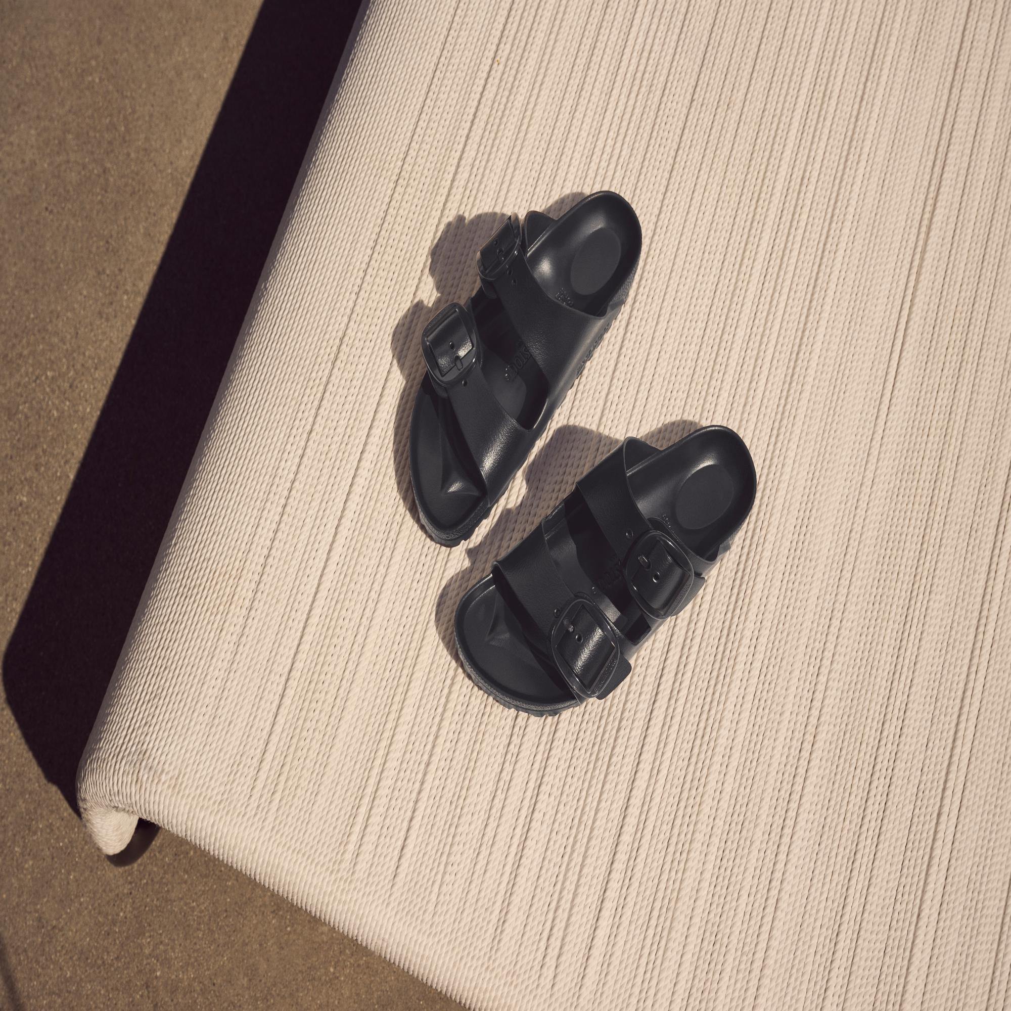 Black Birkenstock Arizona Big Buckle EVA narrow sandals with arch support and buckle straps, placed on a beige textured surface.