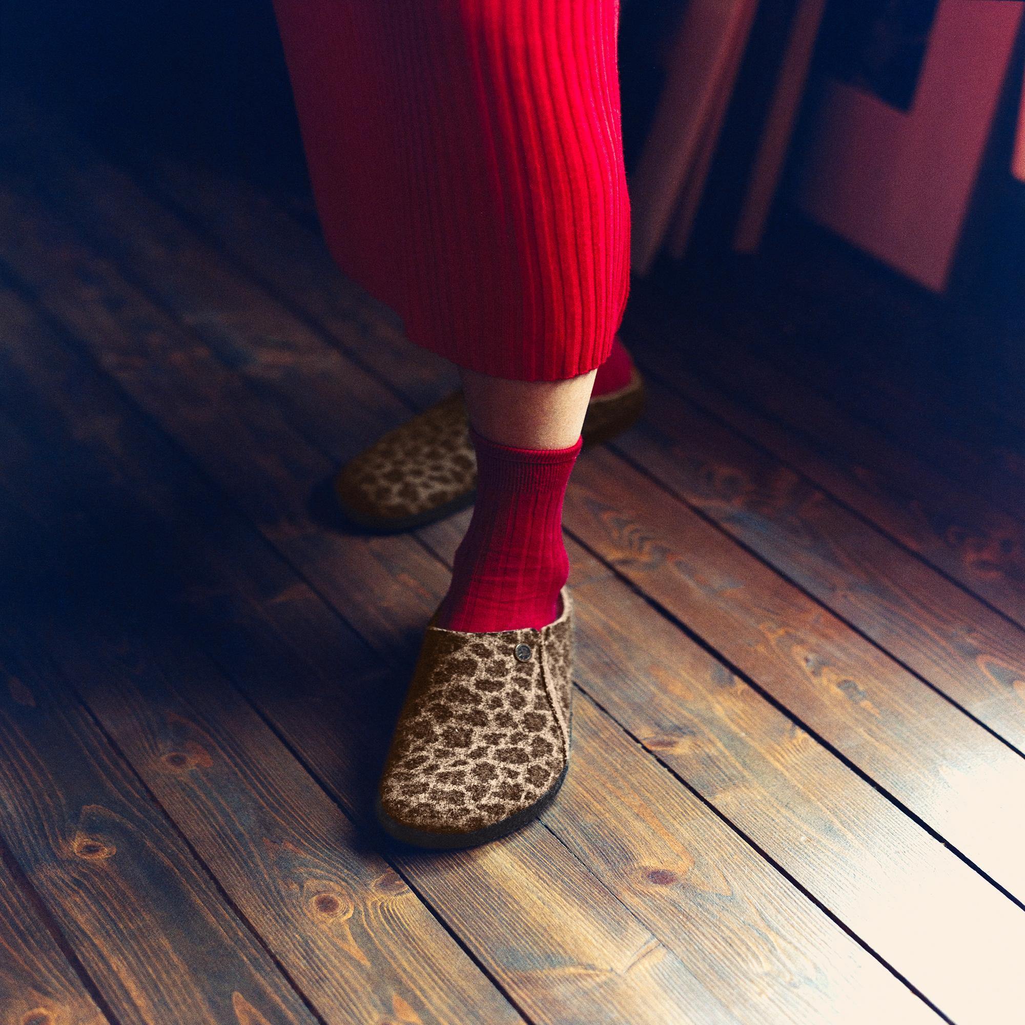 Birkenstock Zermatt Leopard shearling slip-on mules with textile upper, arch support, worn with red socks and red pants on wooden floor.