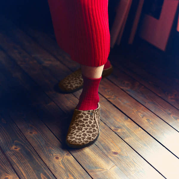 Birkenstock Zermatt Leopard shearling slip-on mules with textile upper, arch support, worn with red socks and red pants on wooden floor.