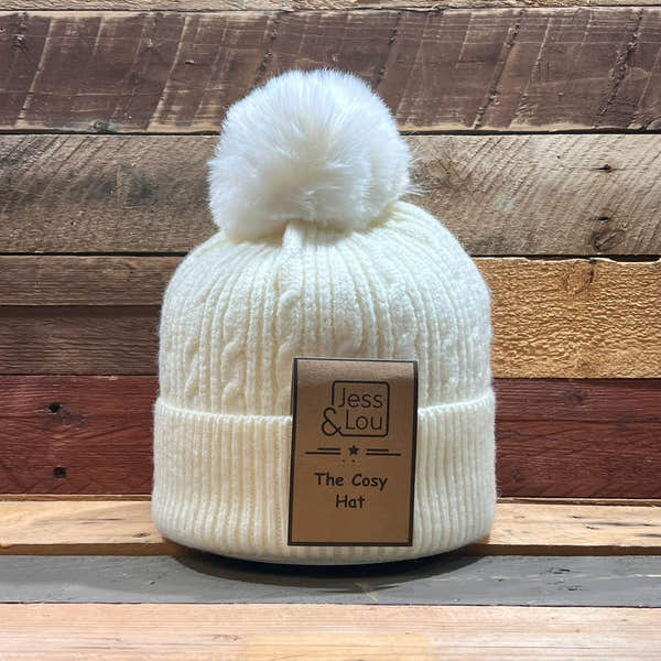 Unisex cream cable knit bobble hat with pom-pom, labeled "Jess & Lou The Cosy Hat" on wooden background.
