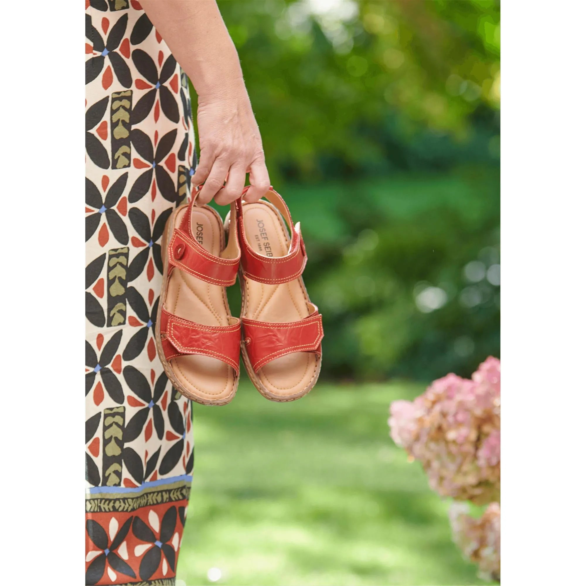 Josef Seibel Debra 58 women's red leather sandals with touch fastening straps, held outdoors against a green background.