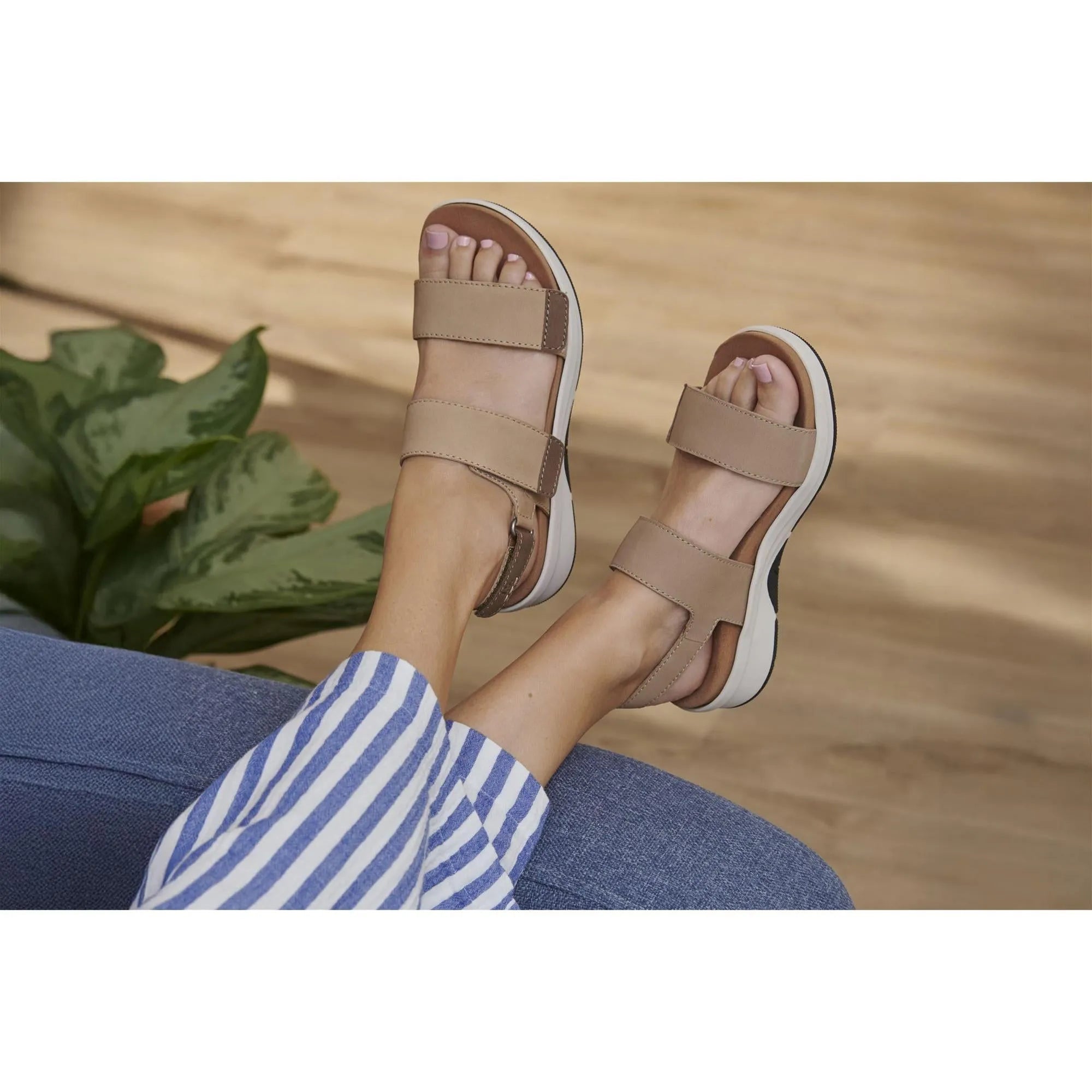 Beige leather touch fastening sandals with straps, open toe, white sole, worn by a person in striped pants, on a wooden floor.