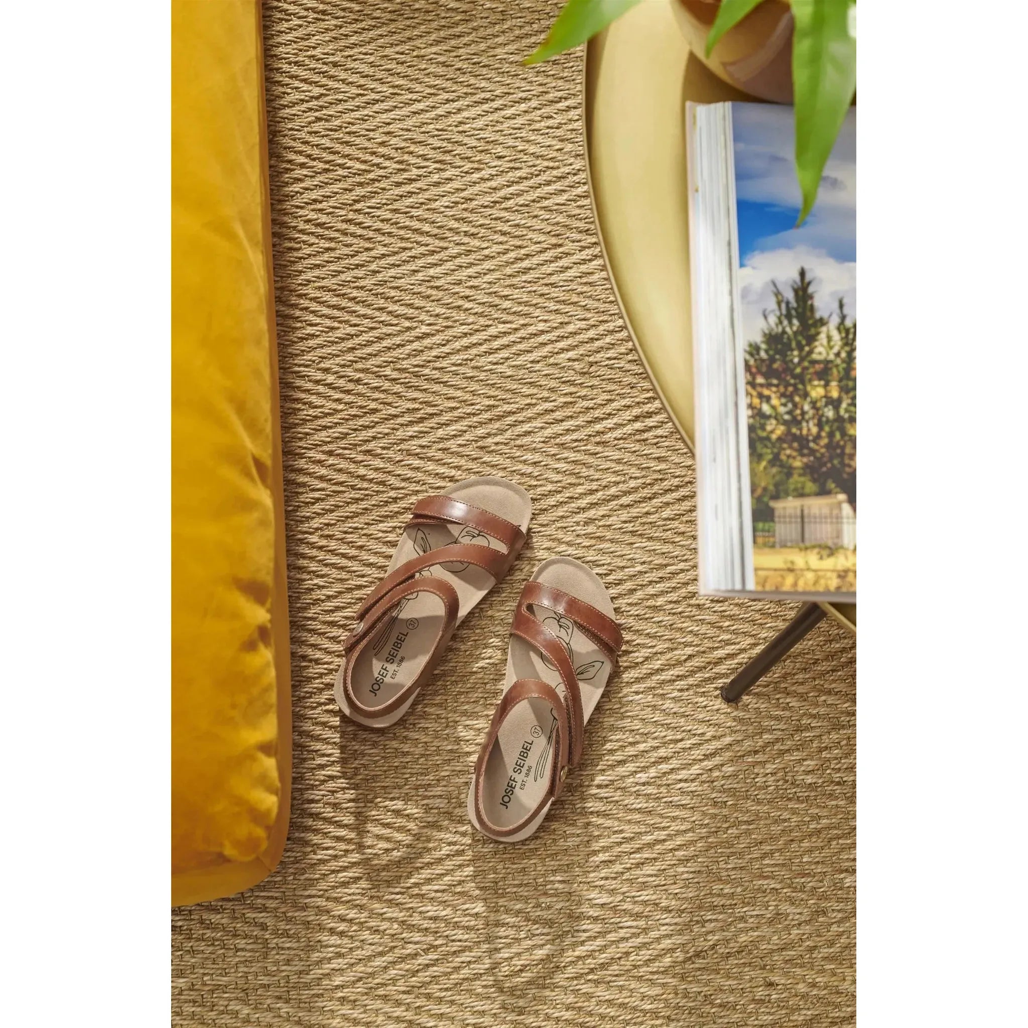 Josef Seibel Tonga 25 women's camel leather sandals on a woven rug, near a yellow table and open magazine.