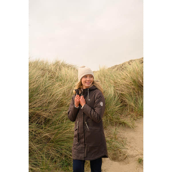 Lighthouse Isobel Cocoa women's padded waterproof coat with a hood, standing outdoors among dunes and tall grass.