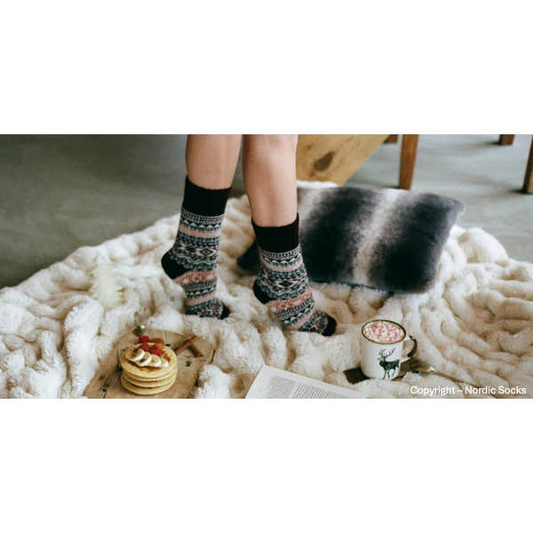Unisex burgundy wool Nordic Socks with intricate patterns, worn on person's legs, on cozy cream faux fur blanket with cookies and hot chocolate.
