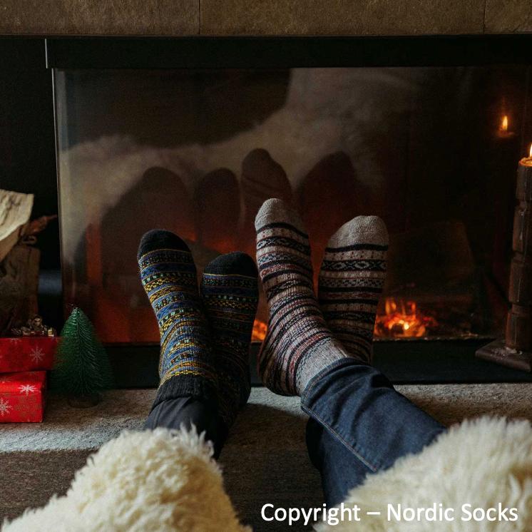 Unisex Arvid wool socks in grey with colorful Nordic patterns, worn by person sitting by a fireplace.