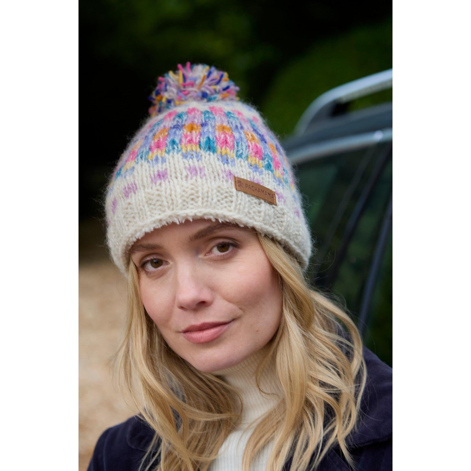 Woman wearing a Pachamama Arundel Bobble Beanie in oatmeal with colorful pom-pom, outdoor setting, casual winter style.
