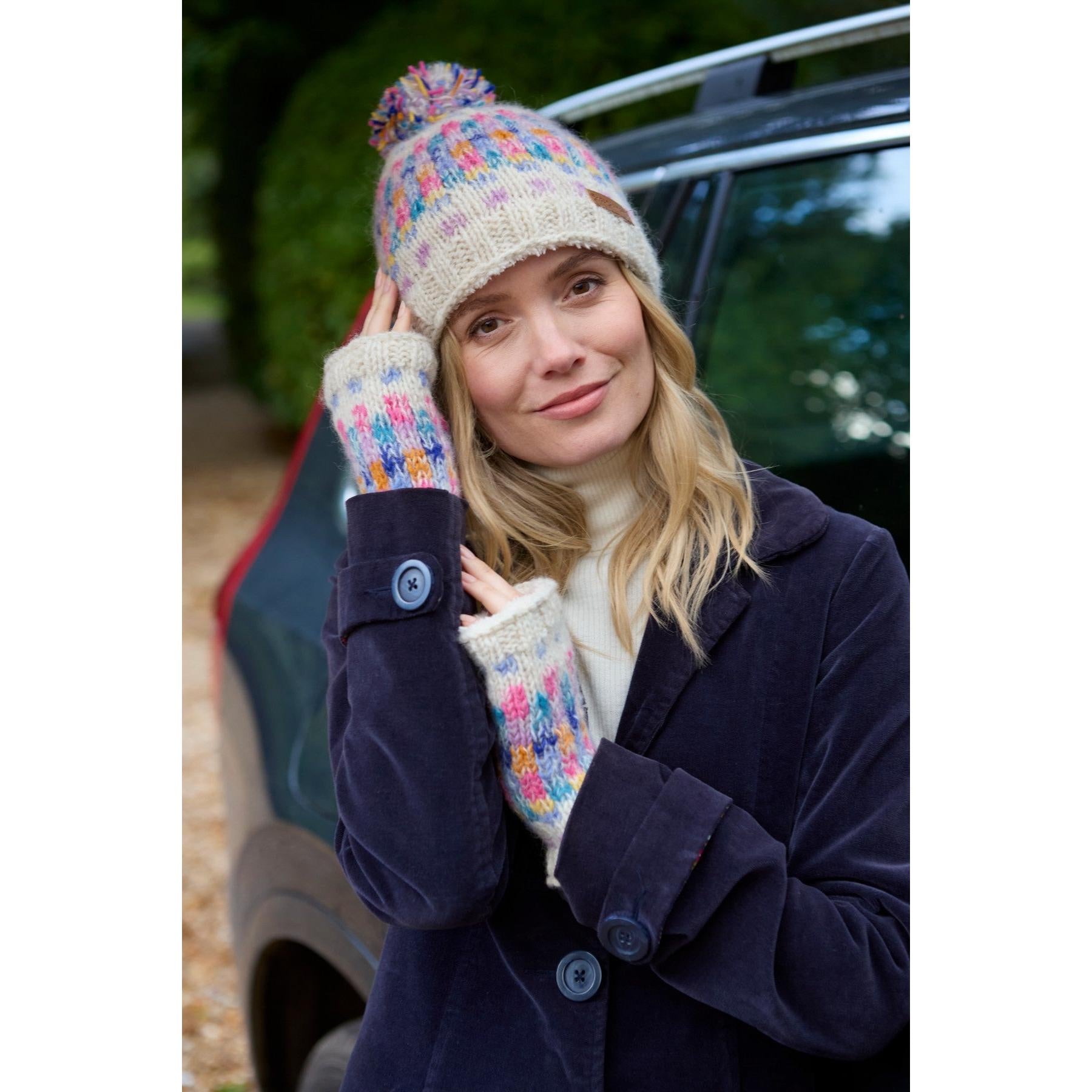 Girl wearing Pachamama Arundel Bobble Beanie and colorful knit mittens outdoors near a car, autumn background, oatmeal-colored beanie.
