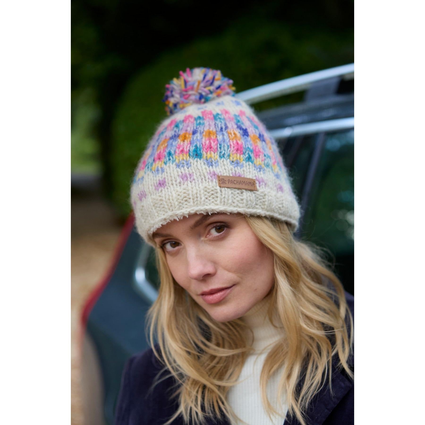 Pachamama Arundel Bobble Beanie in oatmeal with colorful knit pattern and pom-pom, featuring a leather patch, worn by a woman outdoors.