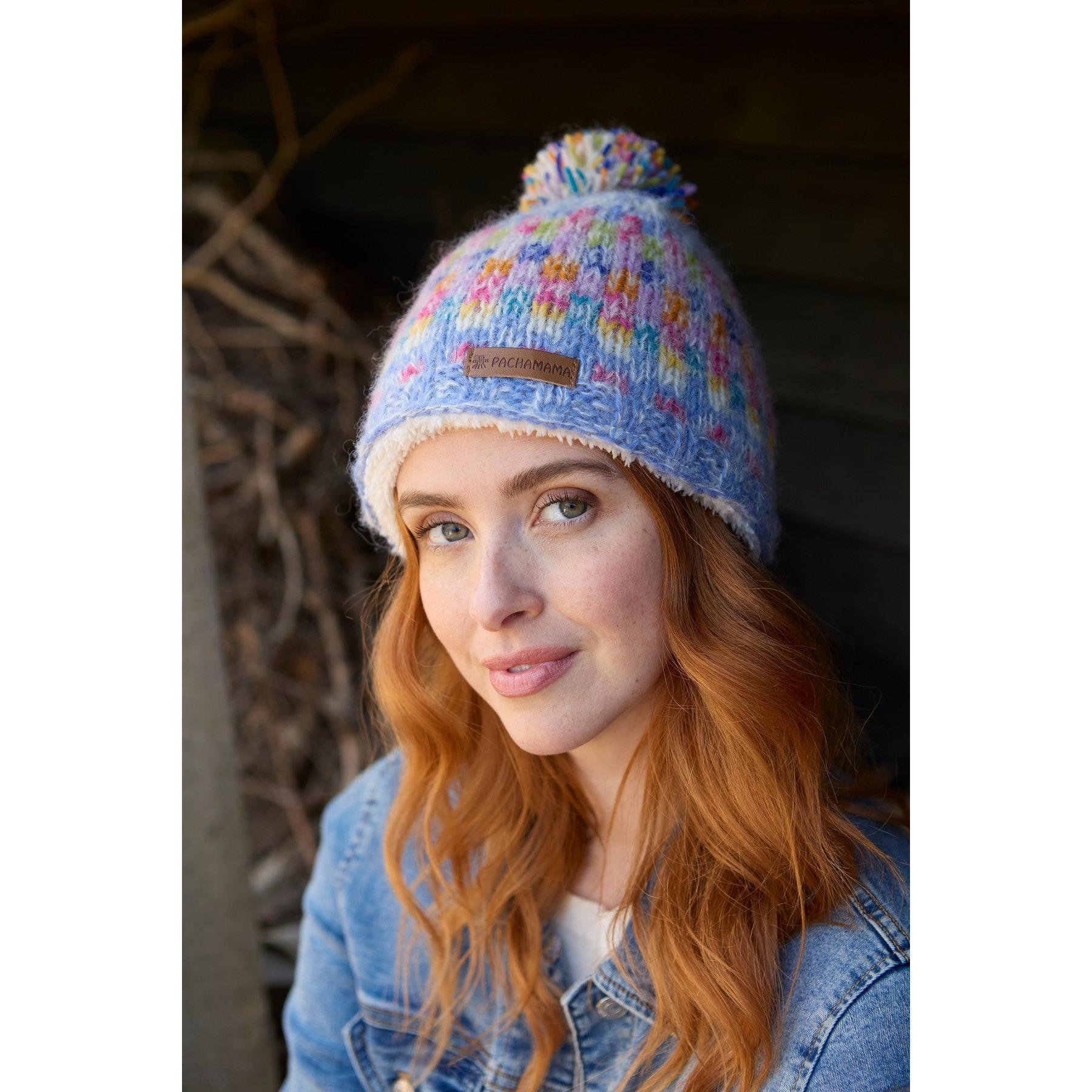 Pachamama Arundel Bobble Beanie Sky, colorful knit winter hat with pom-pom, worn by smiling woman outdoors.