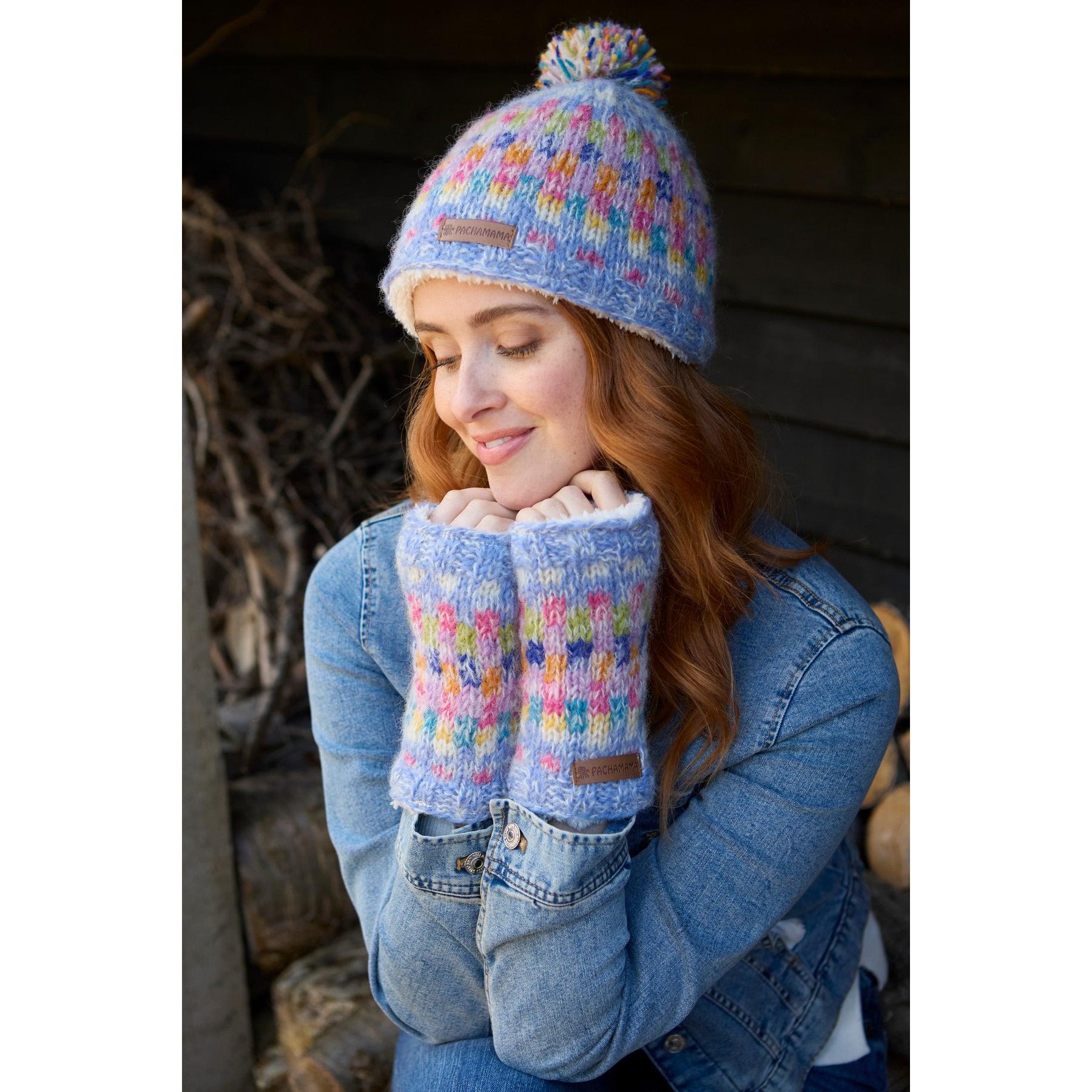 Pachamama Arundel sky-colored bobble beanie and matching fingerless gloves with colorful pattern, worn by smiling woman outdoors.