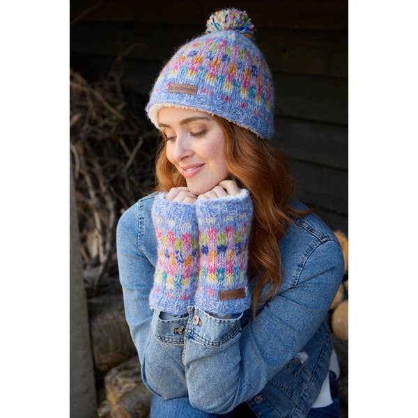 Pachamama Arundel sky-colored bobble beanie and matching fingerless gloves with colorful pattern, worn by smiling woman outdoors.