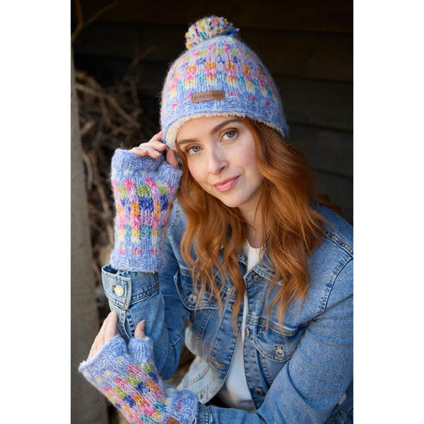 Woman wearing colorful knitted Pachamama Arundel Handwarmer Sky hat and mittens, denim jacket, outdoors.