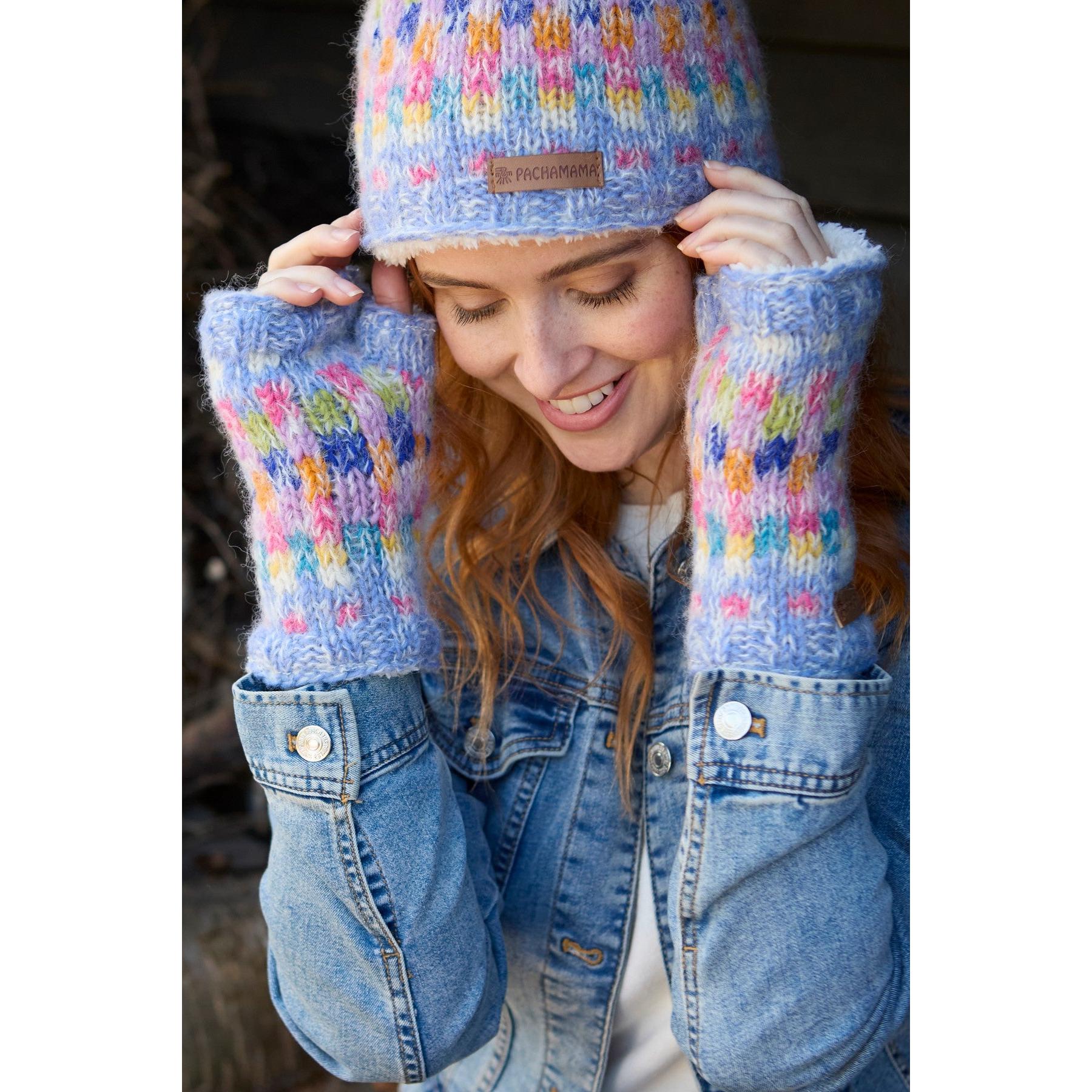 Woman wearing colorful knit Pachamama Arundel Sky hand warmer and beanie, smiling outdoors in denim jacket.