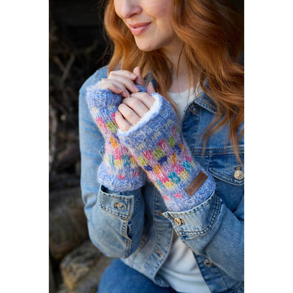 Woman wearing colorful knitted Pachamama Arundel Sky handwarmers, close-up shot, cozy winter accessory, denim jacket, outdoor setting.