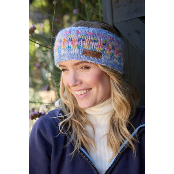 Woman wearing a colorful Pachamama Arundel Sky headband outdoors, smiling, with floral background.