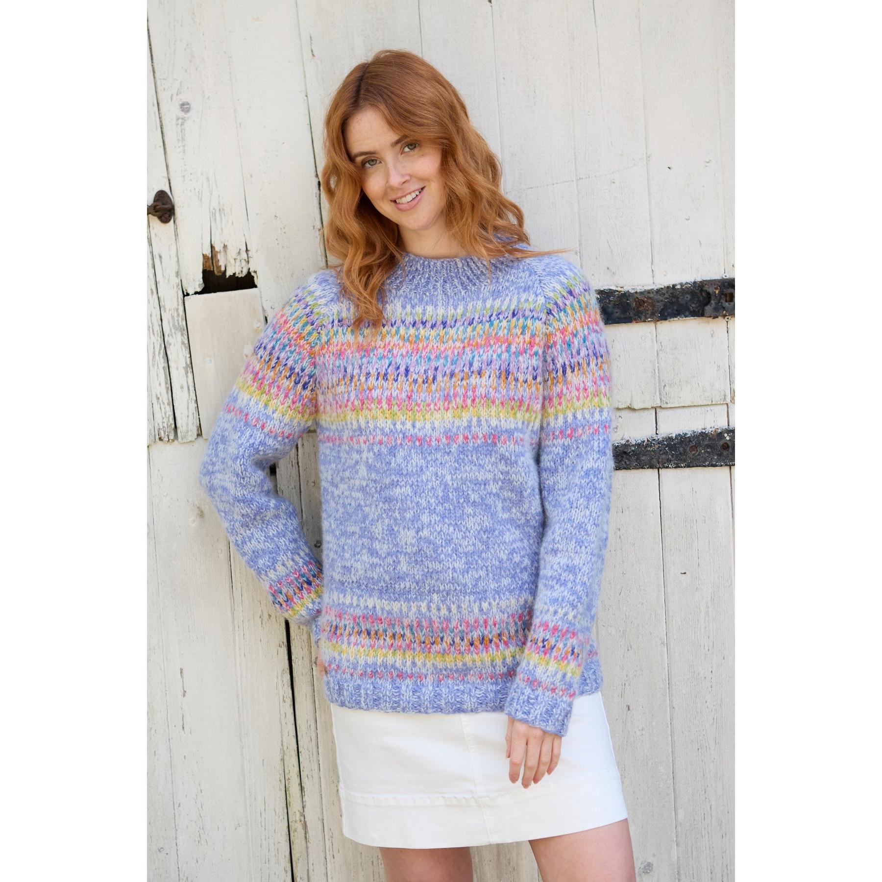Pachamama Arundel Sweater Sky with colorful knit pattern, long sleeves, and relaxed fit, styled with a white skirt, against a rustic wooden background.