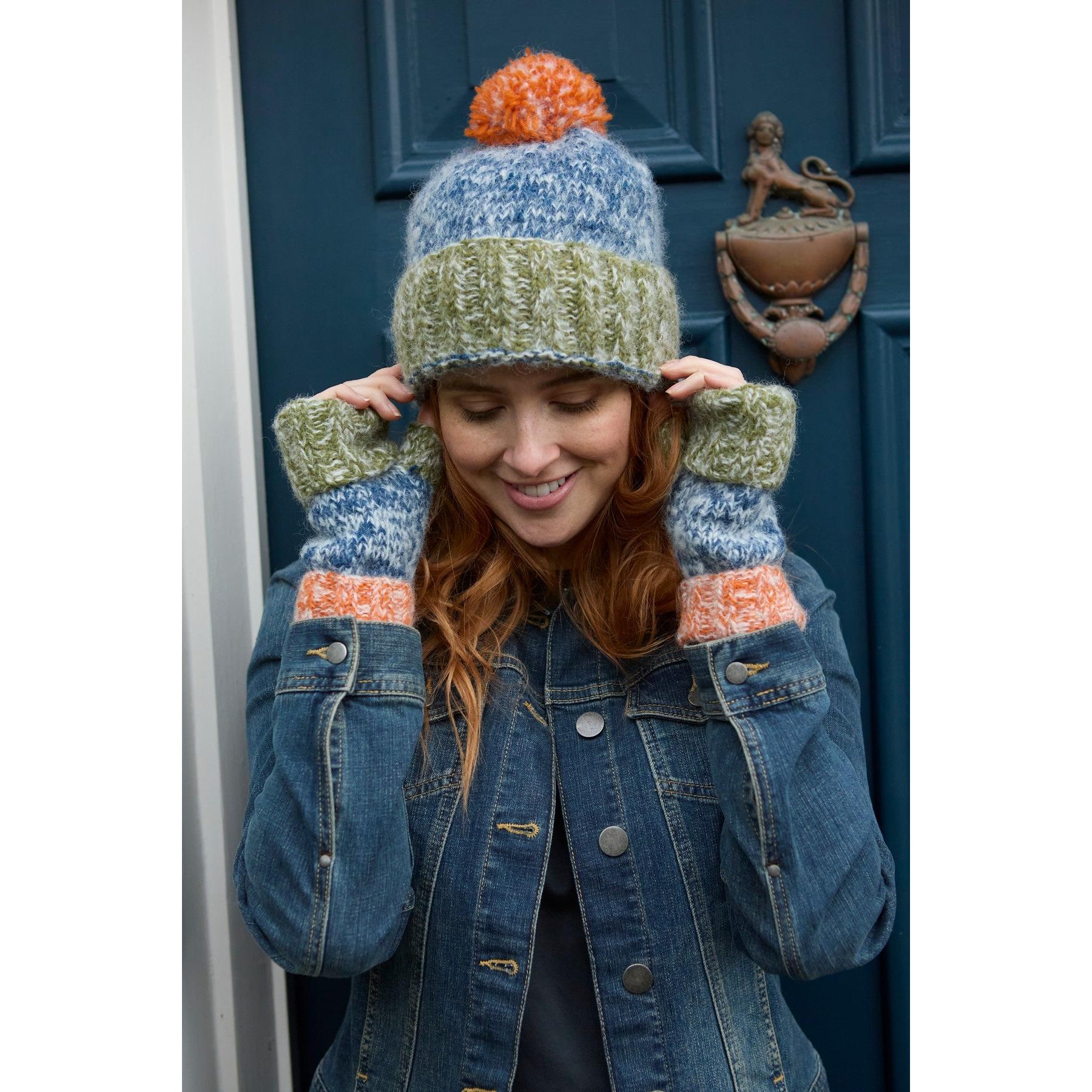 Woman wearing a multicolored knit bobble beanie and fingerless gloves, standing in front of a blue door.
