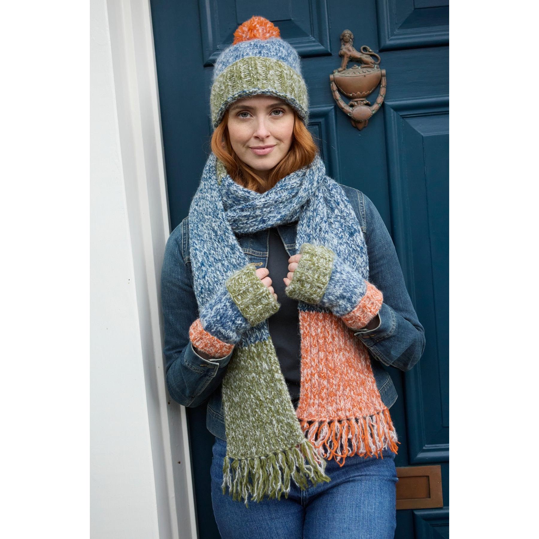Woman wearing blue Pachamama Berlin bobble beanie, matching scarf, and fingerless gloves in front of a blue door.