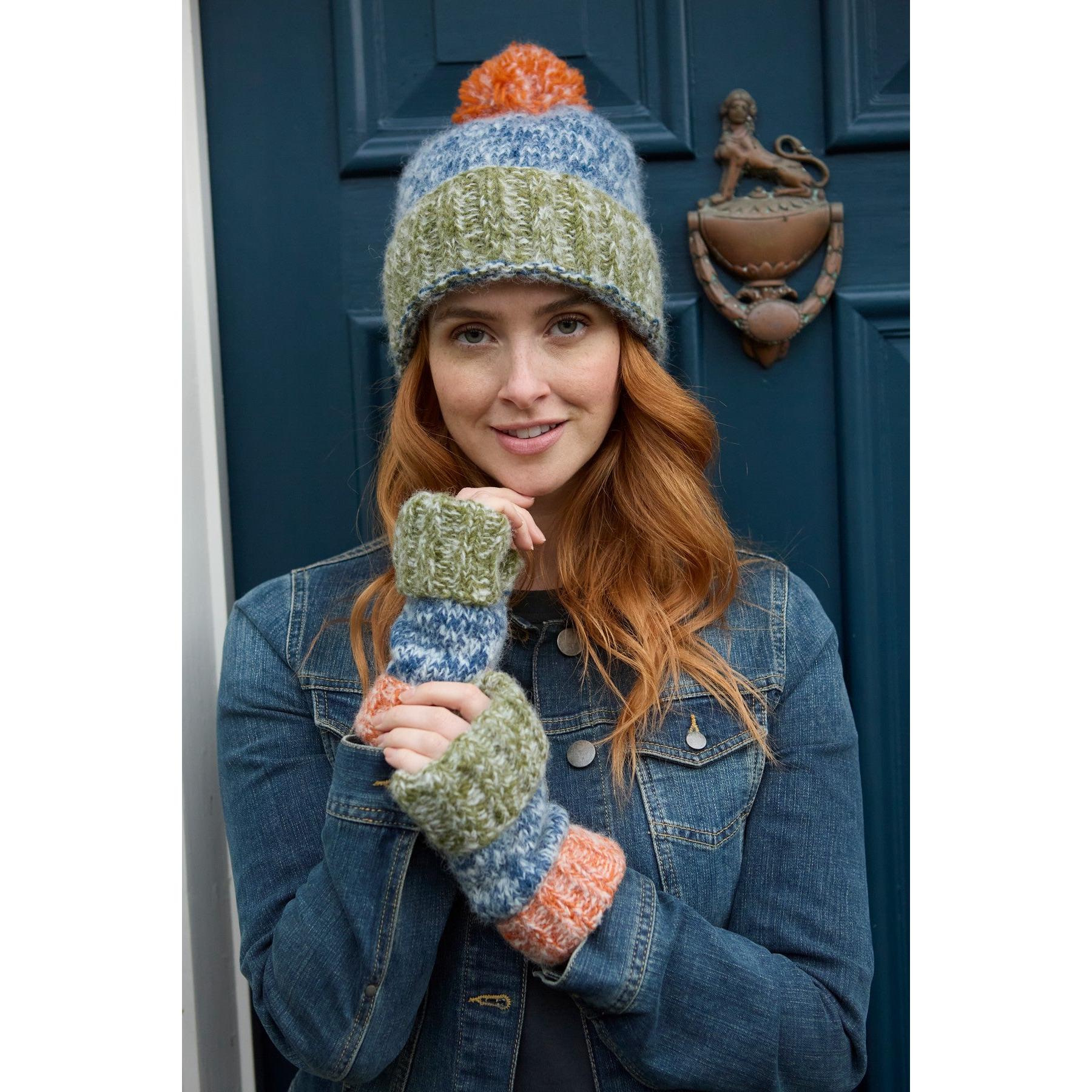 Woman wearing blue, green, orange, and gray knit bobble beanie and fingerless gloves, standing in front of a dark blue door.
