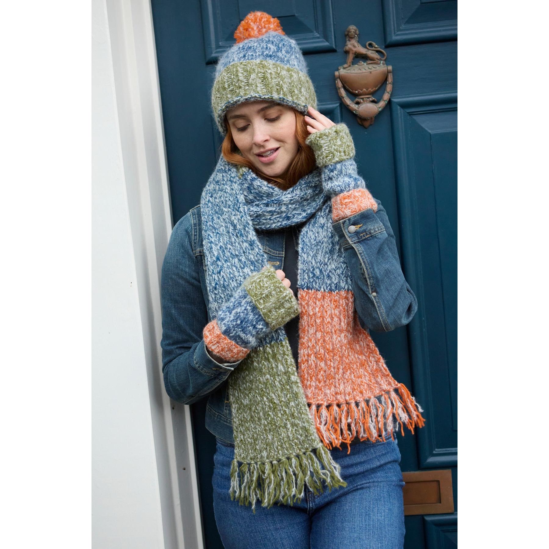 Pachamama Berlin blue, orange, and green bobble beanie, scarf, and gloves set worn by woman outdoors in front of a blue door.