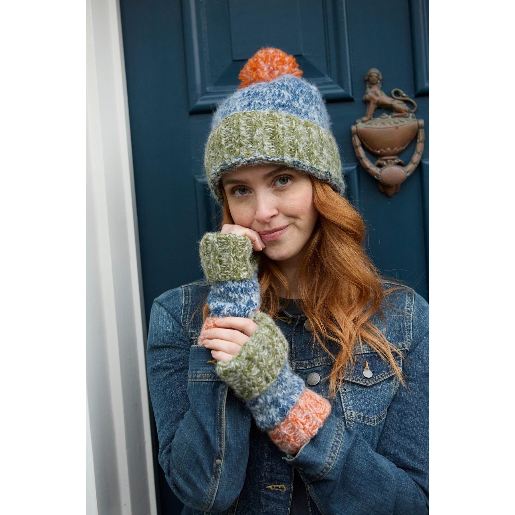Woman wearing blue, multicolored Pachamama Berlin bobble beanie and matching fingerless gloves, standing in front of a blue door.