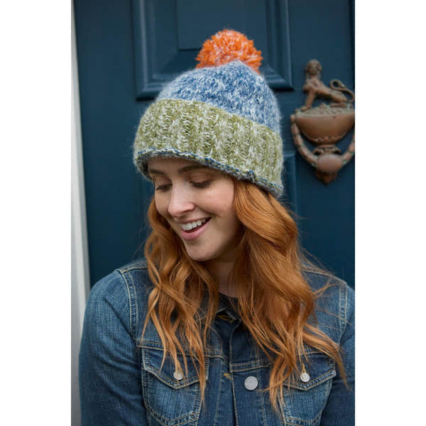 Woman wearing a blue, green, gray, and orange Pachamama Berlin bobble beanie hat with pom-pom, smiling outdoors.
