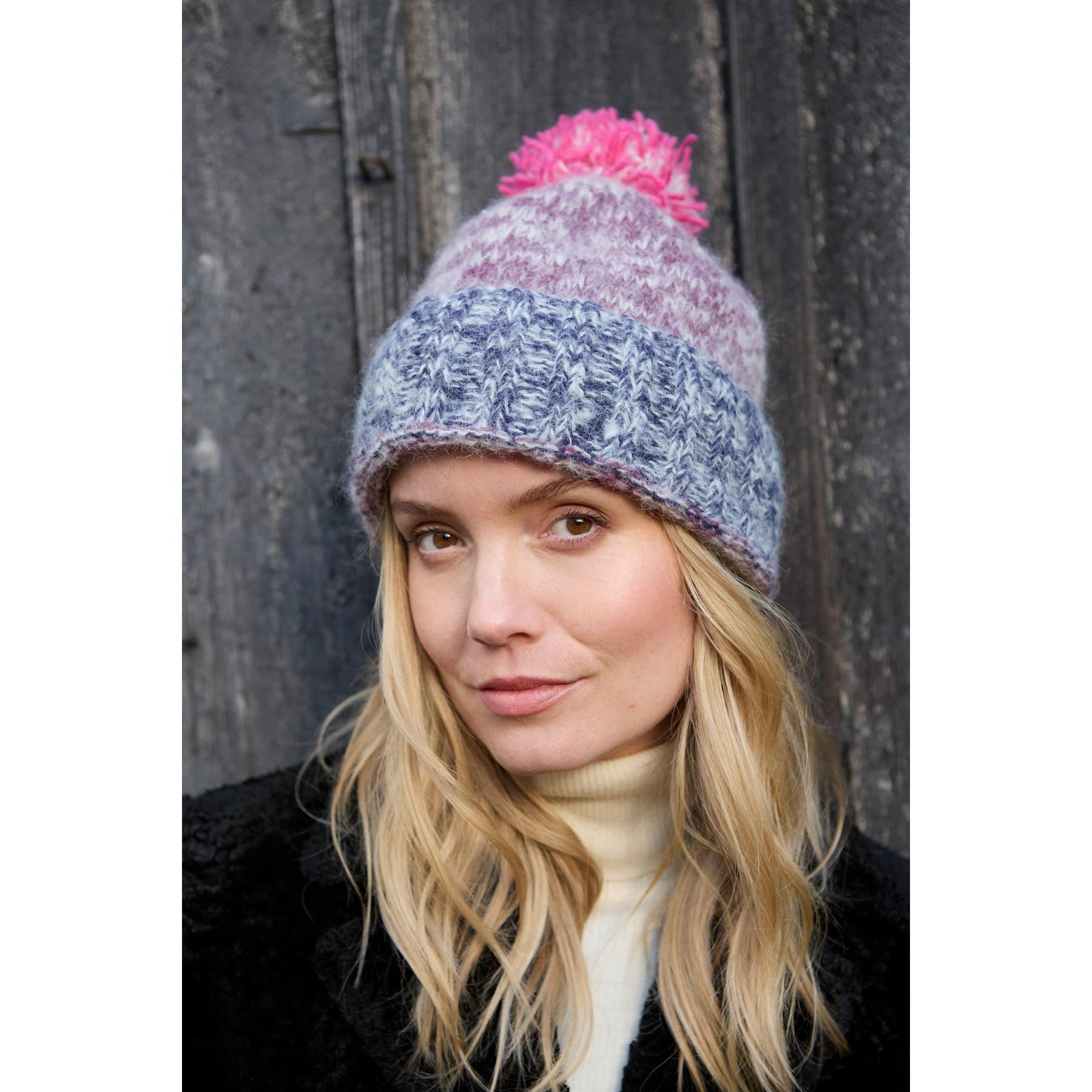 Woman wearing purple Pachamama Berlin bobble beanie with pink pom-pom, knit pattern, and ribbed cuff, outdoors against wooden background.