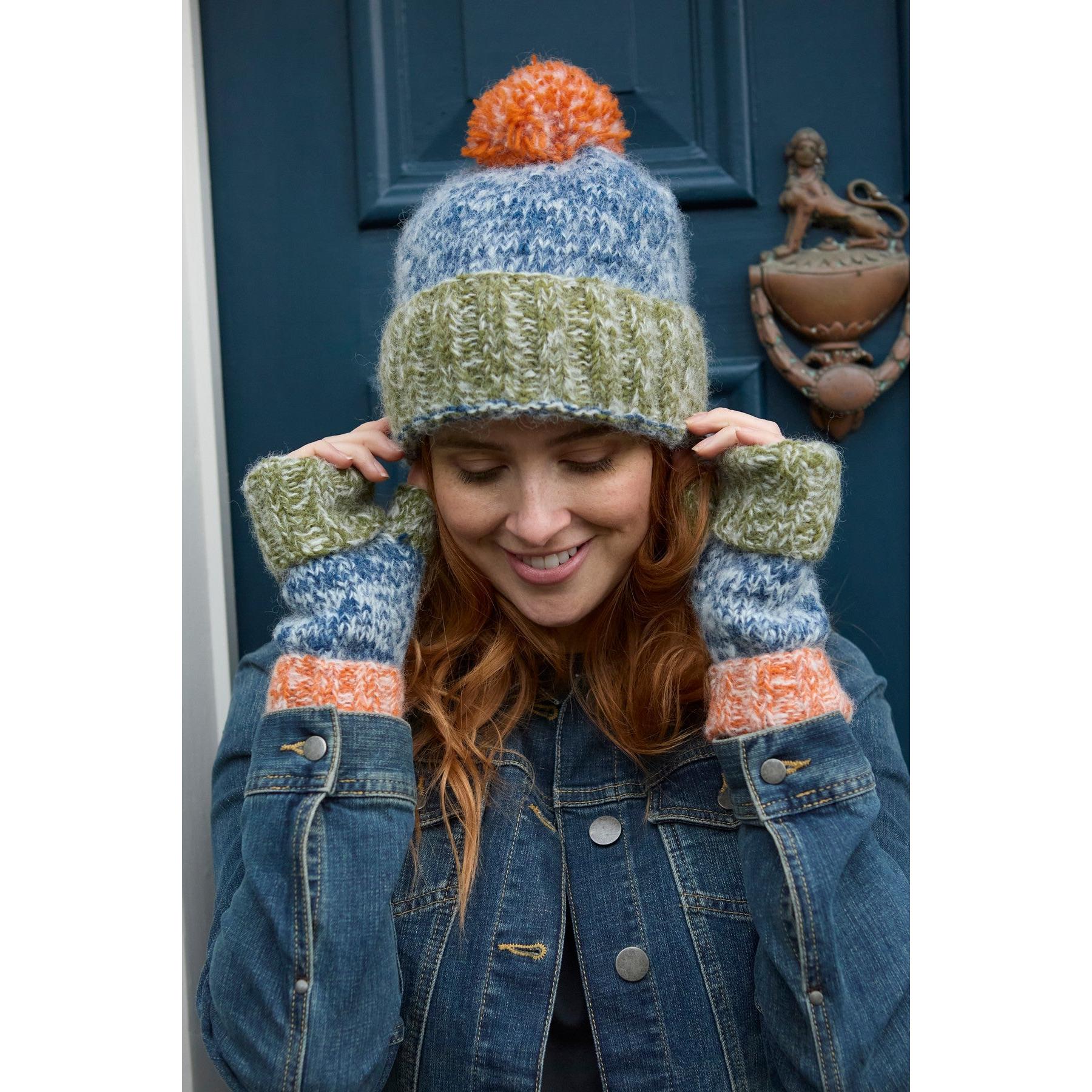 Woman wearing blue, green, orange knit hat and matching mittens, holding the hat's brim, outside on a navy door.