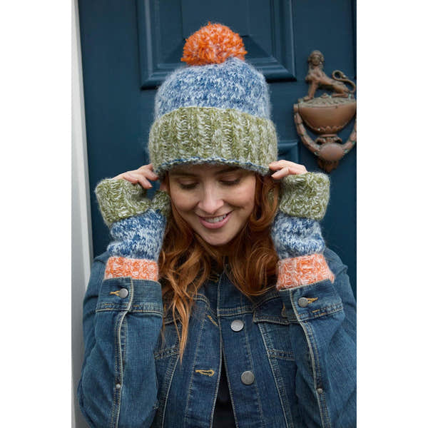 Woman wearing blue, green, orange knit hat and matching mittens, holding the hat's brim, outside on a navy door.