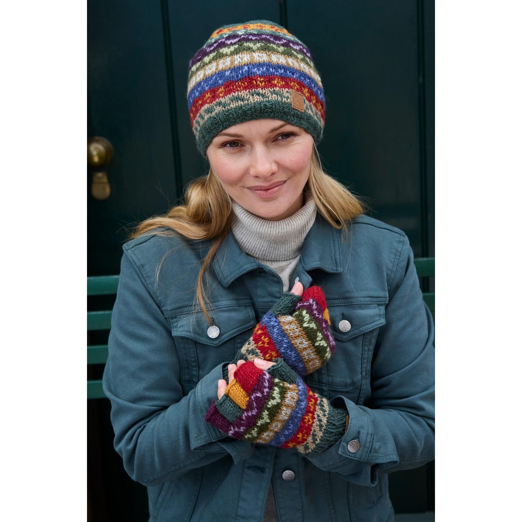 Woman wearing colorful Pachamama Elveden beanie and gloves, denim jacket, smiling outdoors against a dark background.
