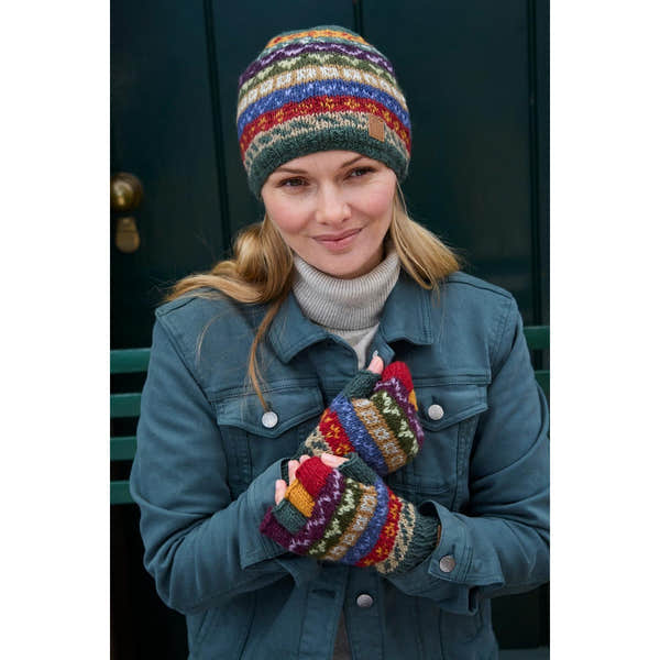 Woman wearing colorful Pachamama Elveden beanie and gloves, denim jacket, smiling outdoors against a dark background.