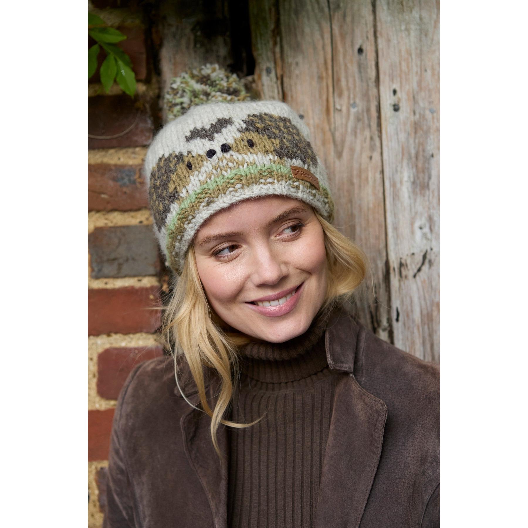 Woman wearing Pachamama Hedgehog bobble beanie with multicolor knit pattern, sitting outdoors near a wooden wall.