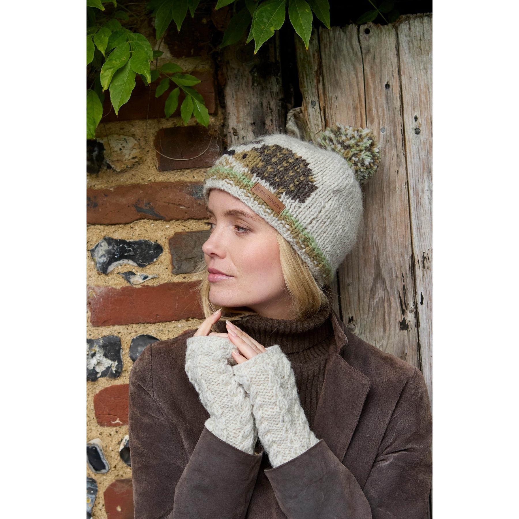 Woman wearing a beige Pachamama Hedgehog Bobble Beanie with a pom-pom, standing outdoors against a brick and wood wall.