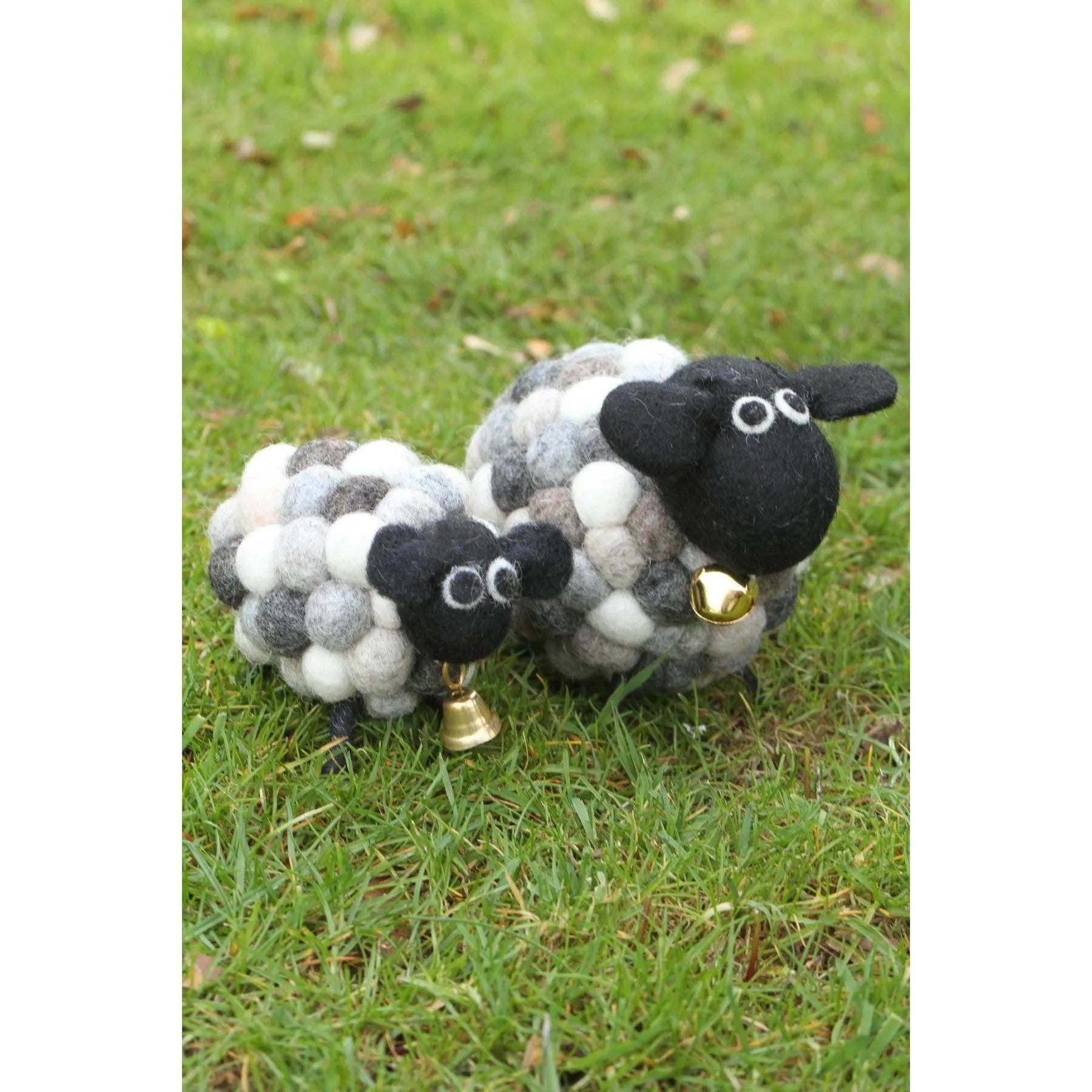 Felted wool lamb and sheep plush toys with black faces, white bodies, googly eyes, and gold bells on grass.