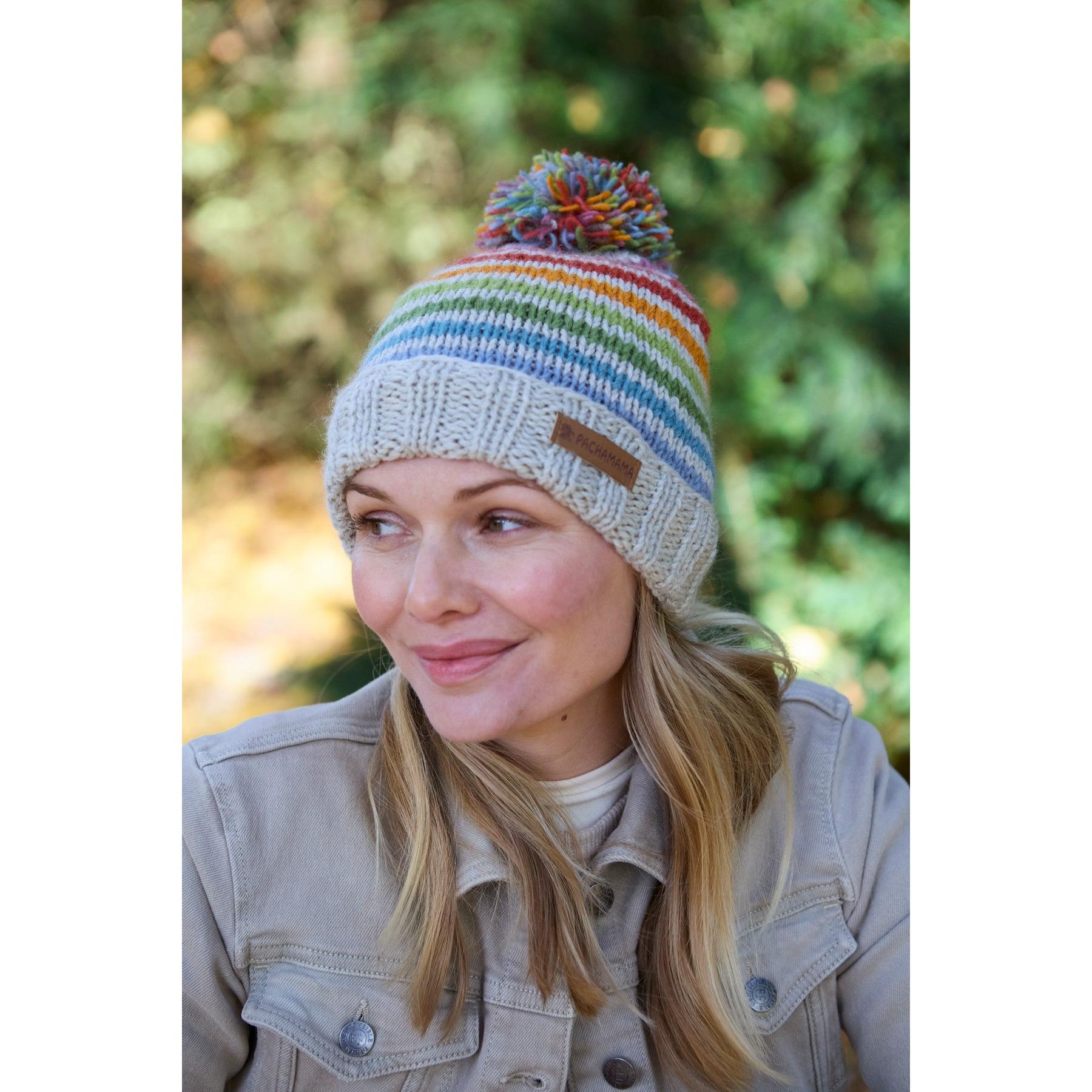 Pachamama Hoxton Stripe Bobble Beanie in pure merino wool with colorful stripes and a pom-pom, worn by a smiling woman outdoors.