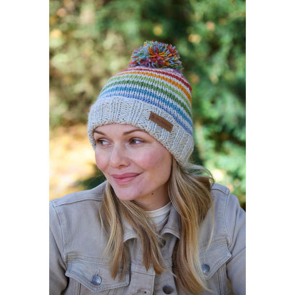 Pachamama Hoxton Stripe Bobble Beanie in pure merino wool with colorful stripes and a pom-pom, worn by a smiling woman outdoors.