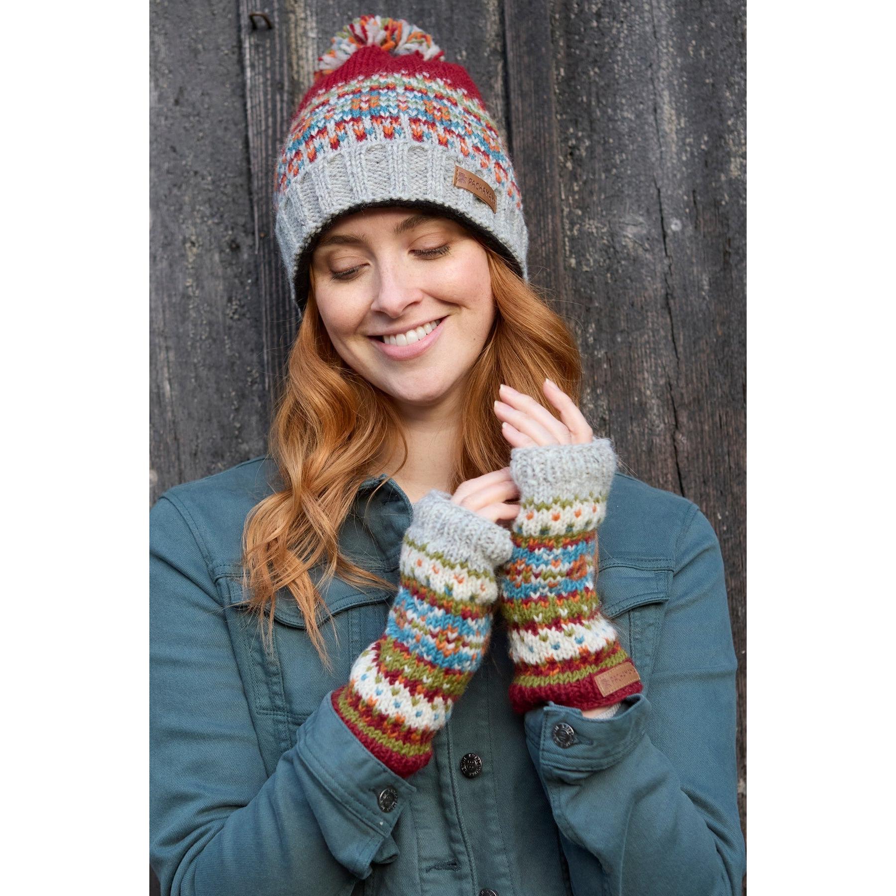 Pachamama Iona Handwarmer Burgundy, colorful knit mittens and cozy hat, woman with red hair smiling outdoors against wooden background.