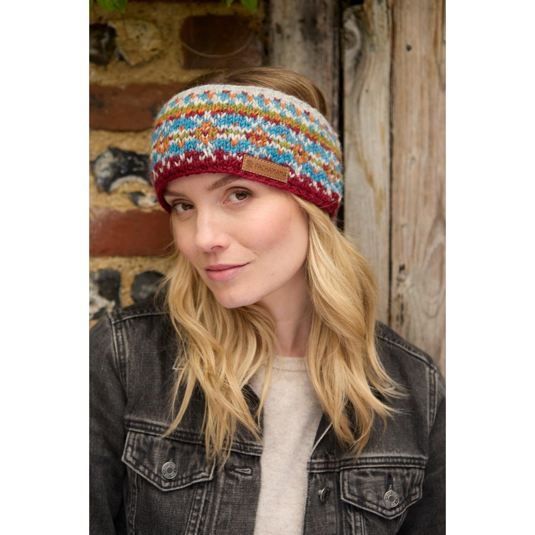 Women wearing burgundy Pachamama Iona knitted headband with multicolored pattern, outdoors against wooden wall.