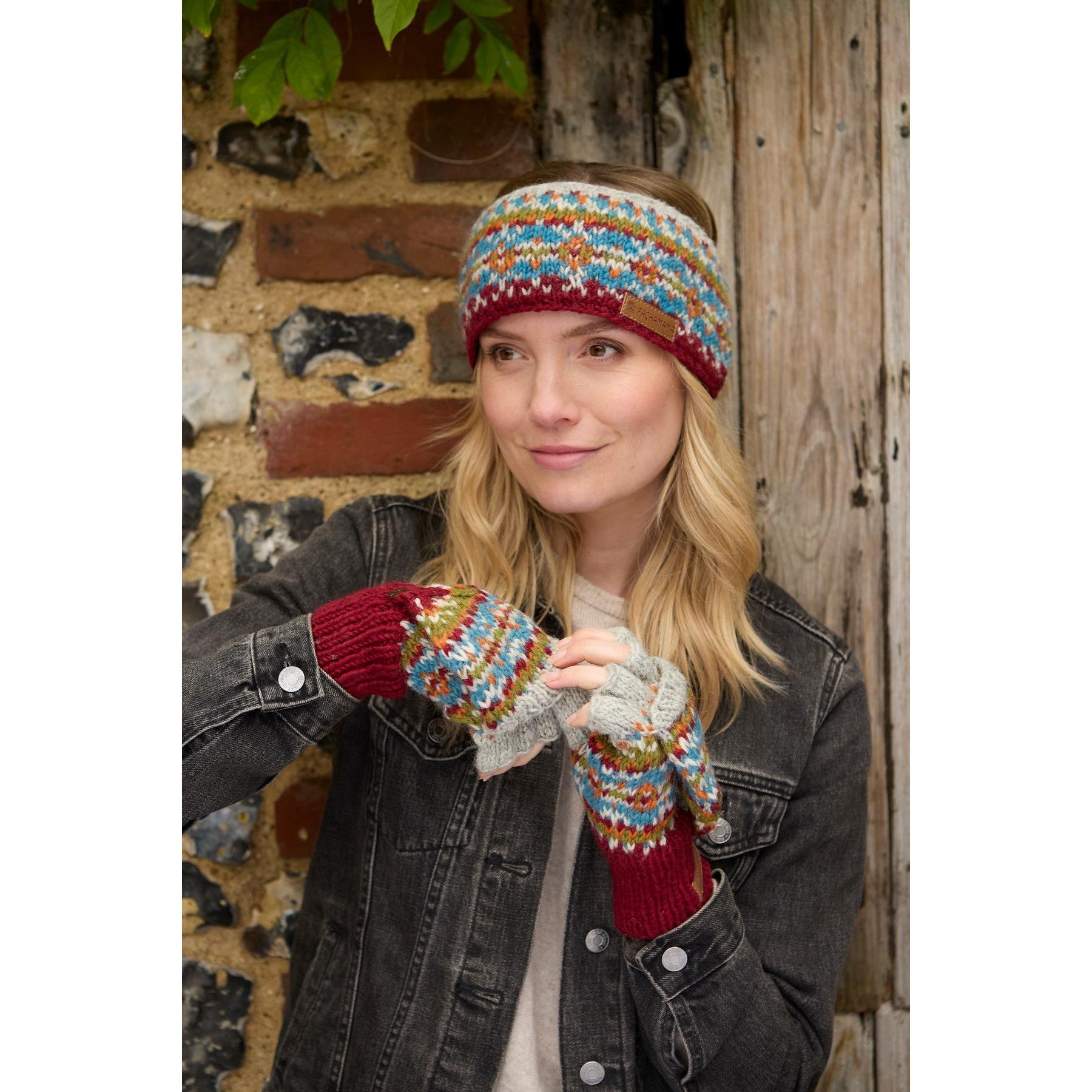 Woman wearing burgundy Pachamama Iona knit headband and multicolored patterned gloves, outdoors with a rustic brick and wooden background.