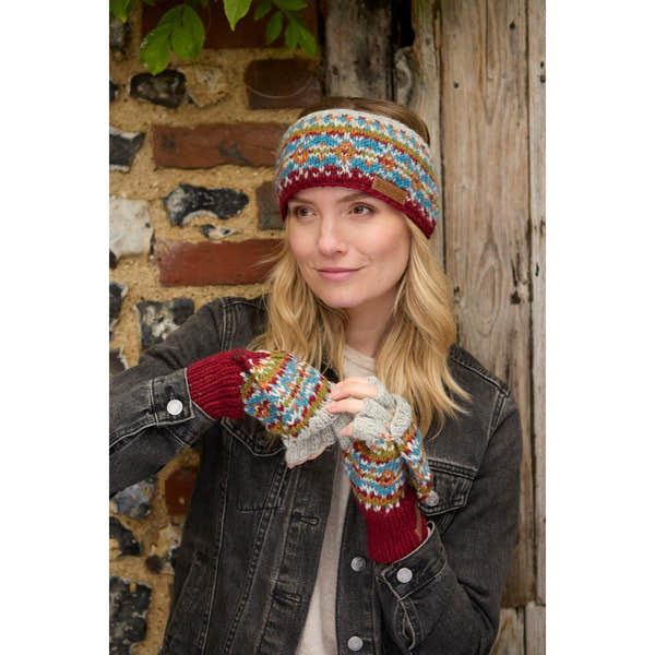 Woman wearing burgundy Pachamama Iona knit headband and multicolored patterned gloves, outdoors with a rustic brick and wooden background.
