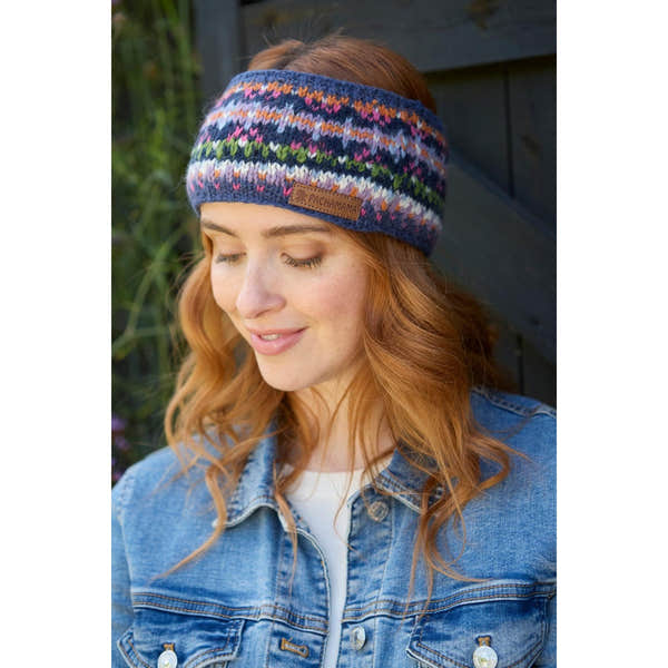 Pachamama Iona Headband Denim with colorful knit pattern and leather logo patch, worn by a smiling woman with red hair outdoors.