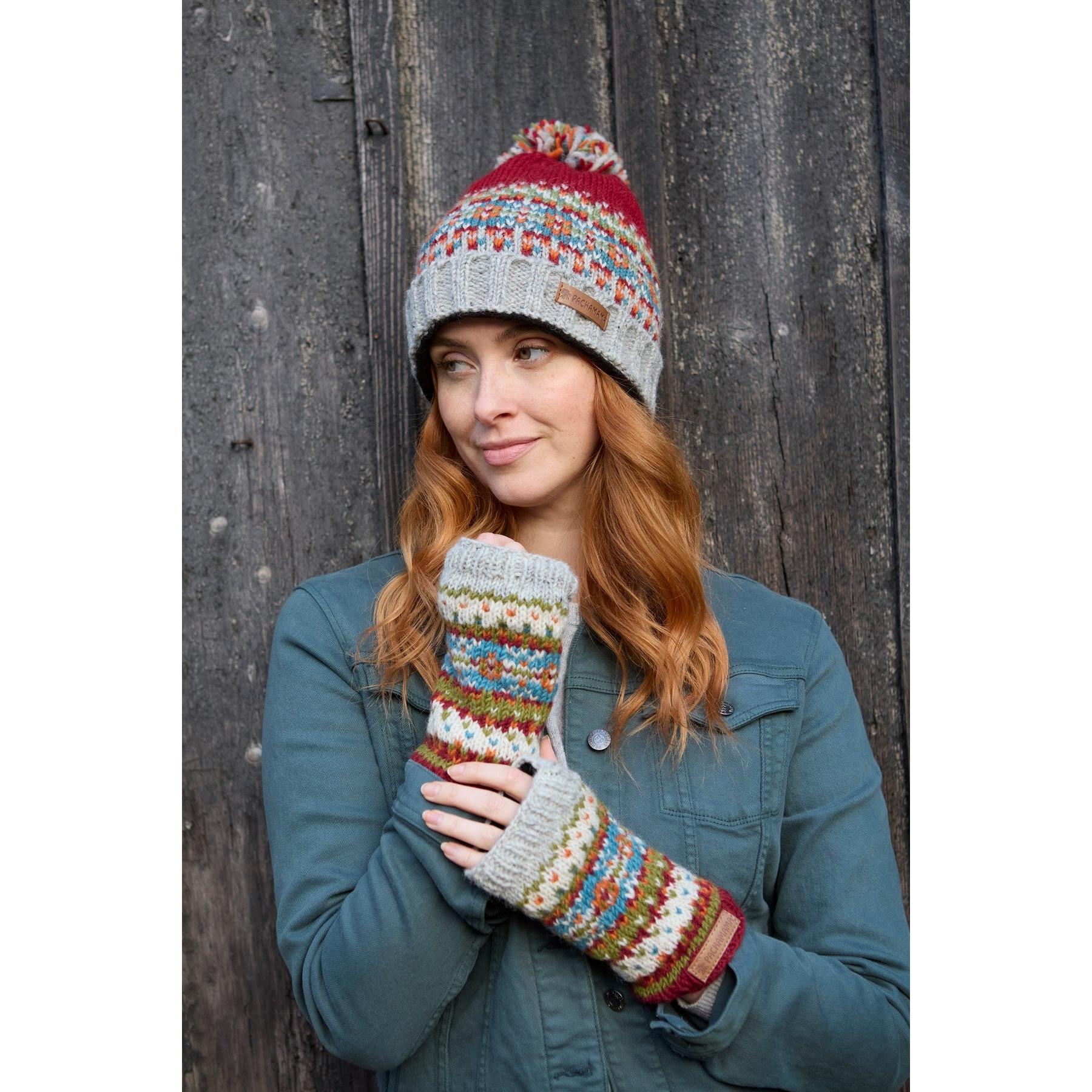Woman wearing burgundy Pachamama Iona Roll Back Bobble Beanie and colorful knitted gloves, outdoor wood background.