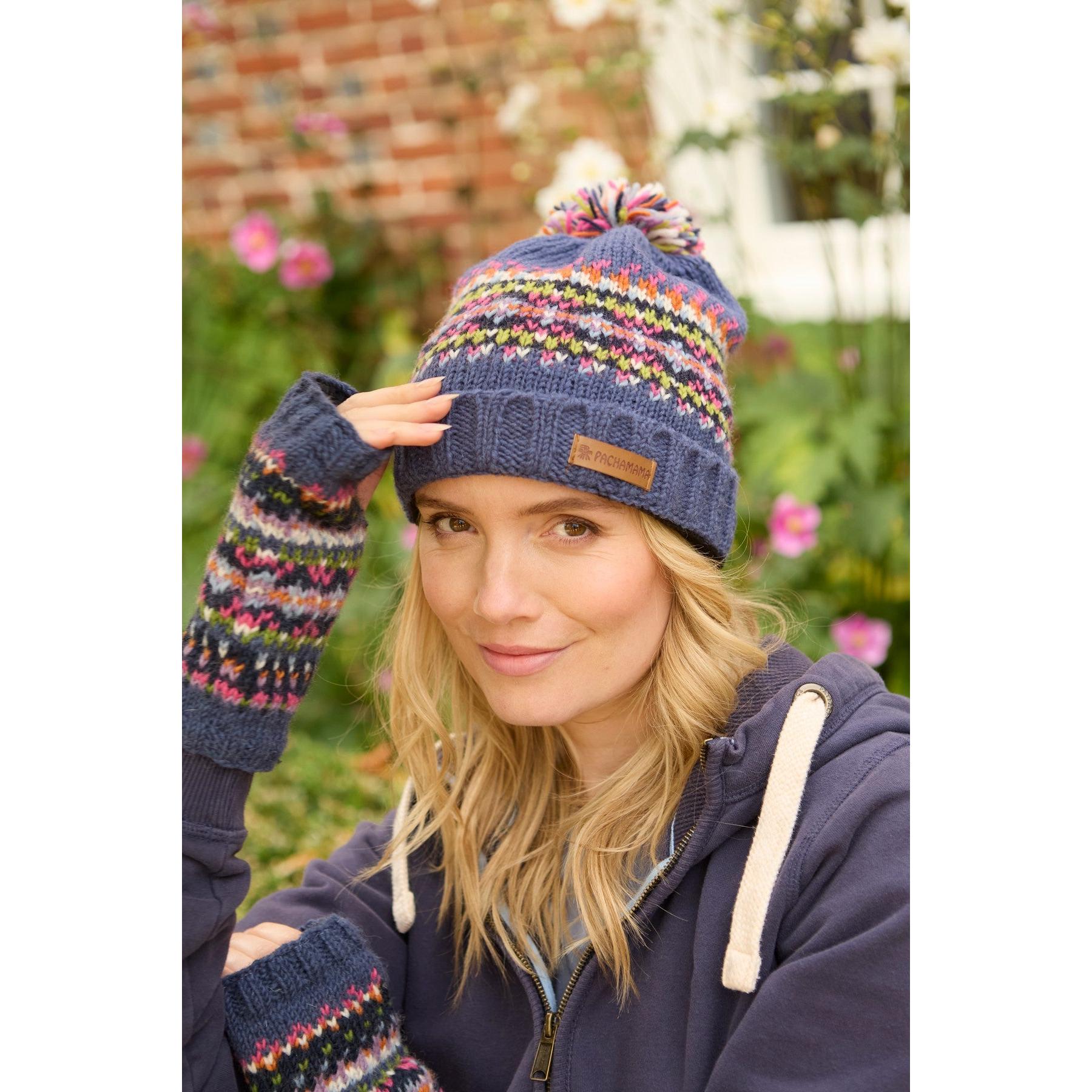 Woman wearing Pachamama Iona Roll Back Bobble Beanie and matching gloves in denim with colorful knit pattern, outdoor garden background.