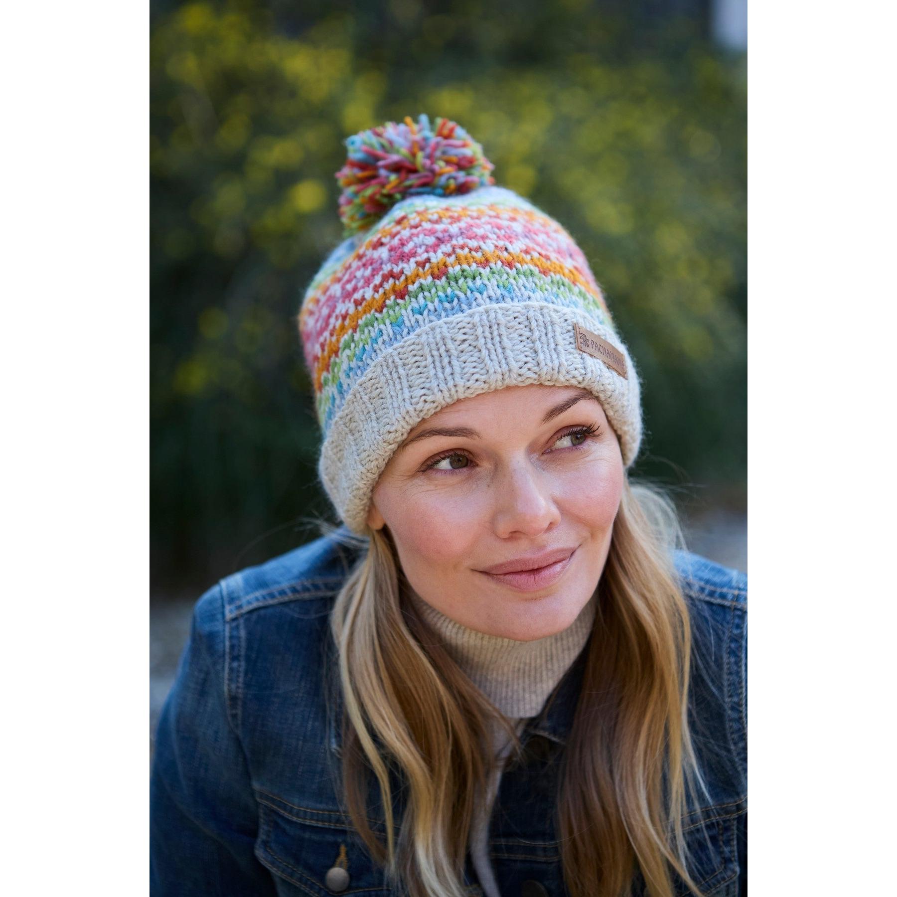 Pachamama Killarney Bobble Beanie in oatmeal with colorful, striped pattern and pompom, made from pure Merino wool, worn by a woman outdoors.