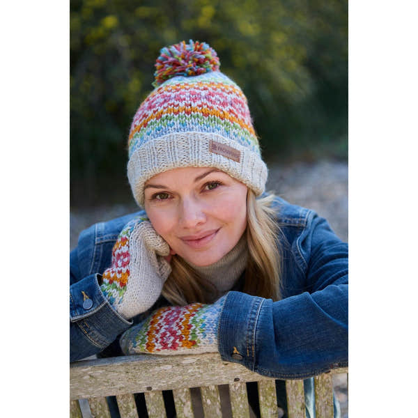 Pachamama Killarney Bobble Beanie in oatmeal with colorful stripes and pom-pom, made of pure merino wool, worn by a smiling woman outdoors.